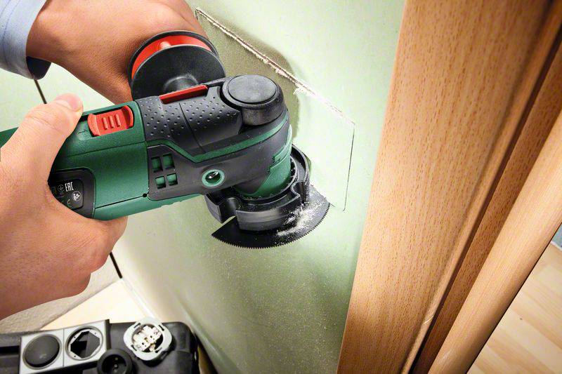 A person is cutting a rectangular hole in a green wall next to a wooden door frame using an electric multi-tool.