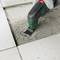 An electric tool with a blade removes grout between tiles. A hand wearing a work glove firmly grips the tool.