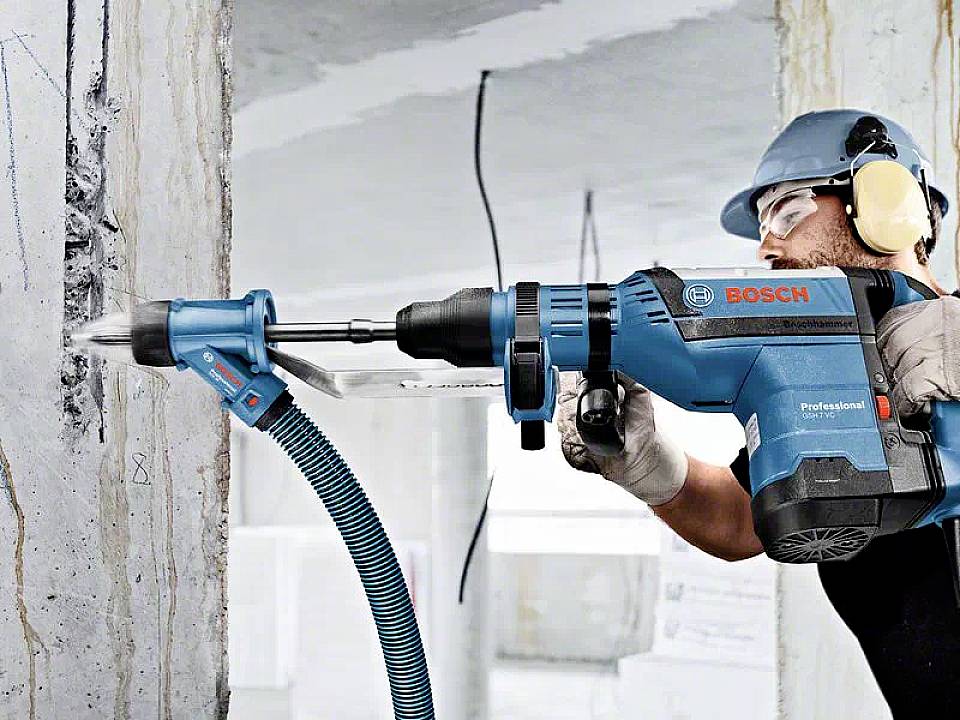 'Worker wearing safety glasses and ear protection uses a Bosch hammer drill on a concrete wall, with dust being extracted through a hose.'