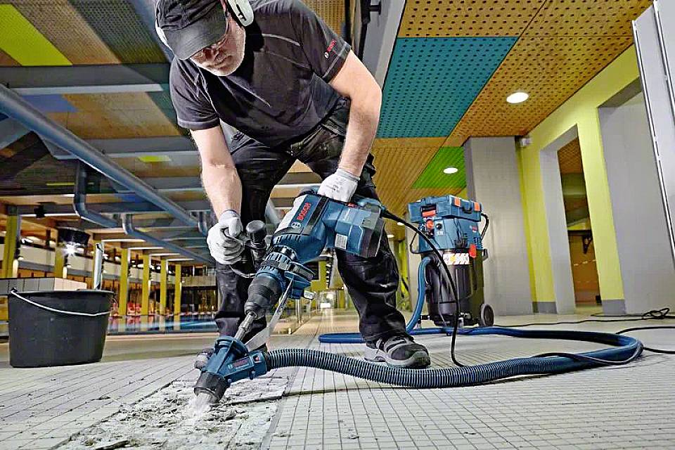 A worker wearing protective clothing is using an electric hammer tool to remove tiles from a floor in a modern building environment.