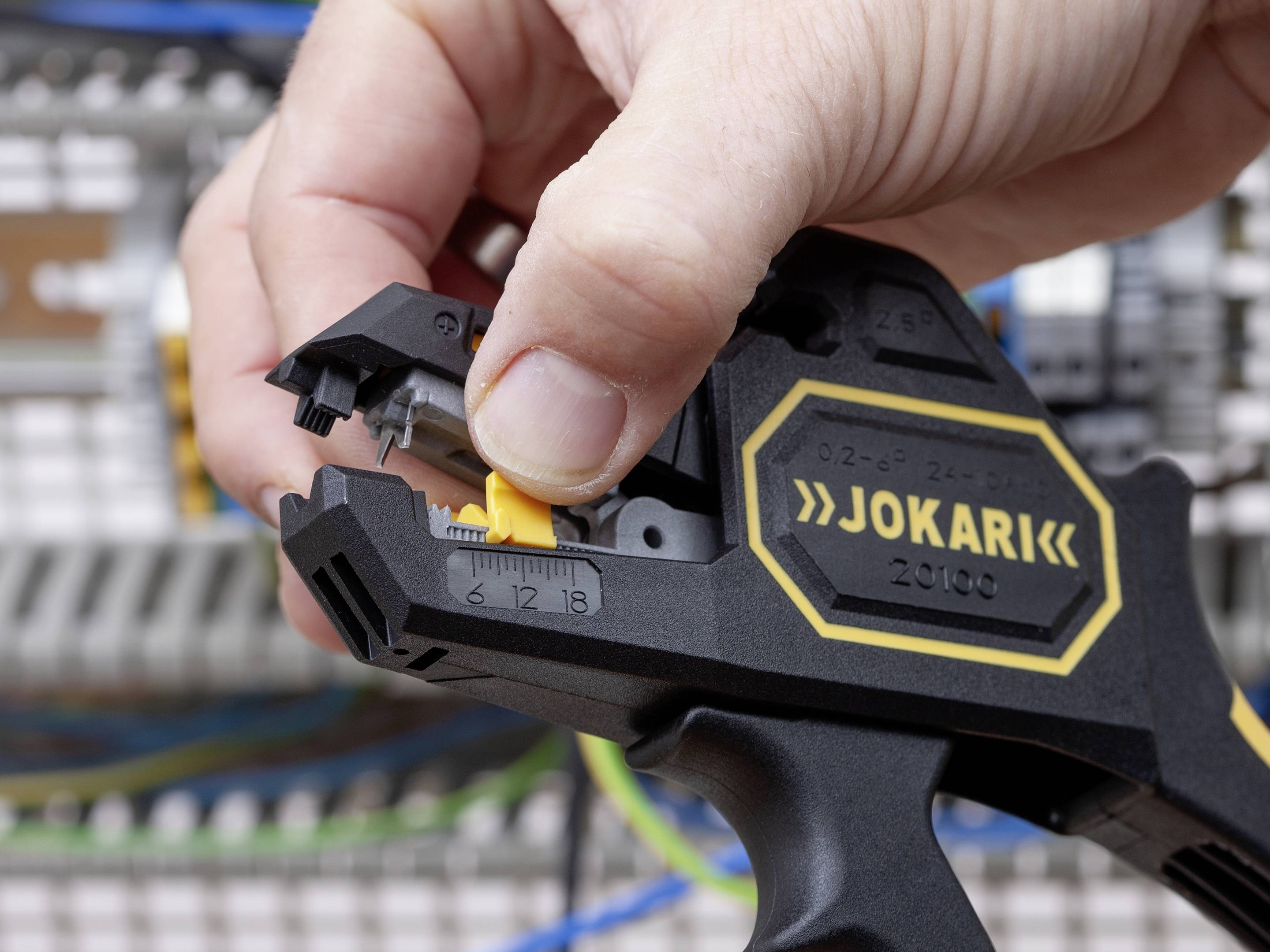 A hand is operating a cable stripping tool, with wires and switchboards visible in the background.