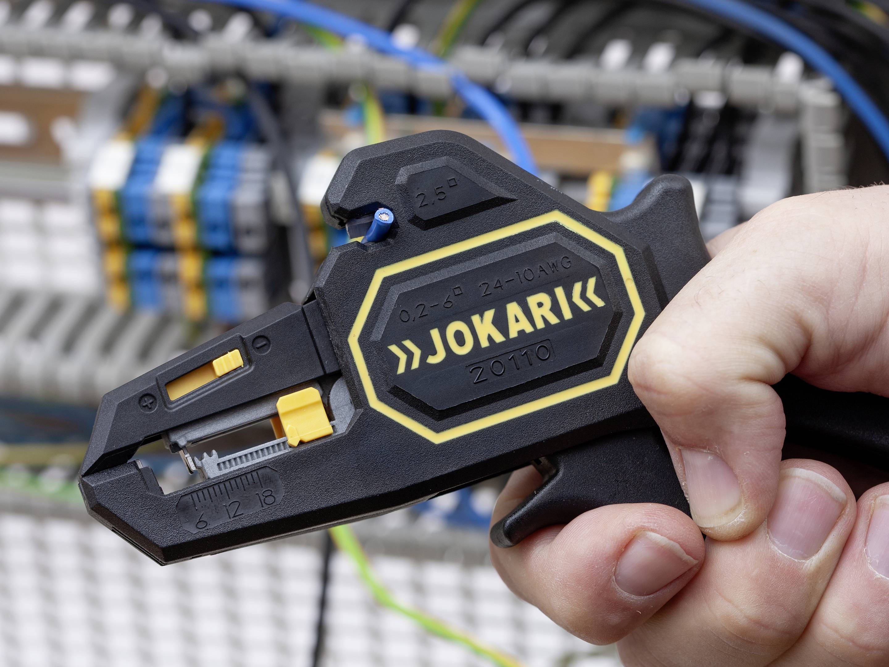 A hand holds a wire stripping tool in front of an electrical cabinet with wired connections in the background.