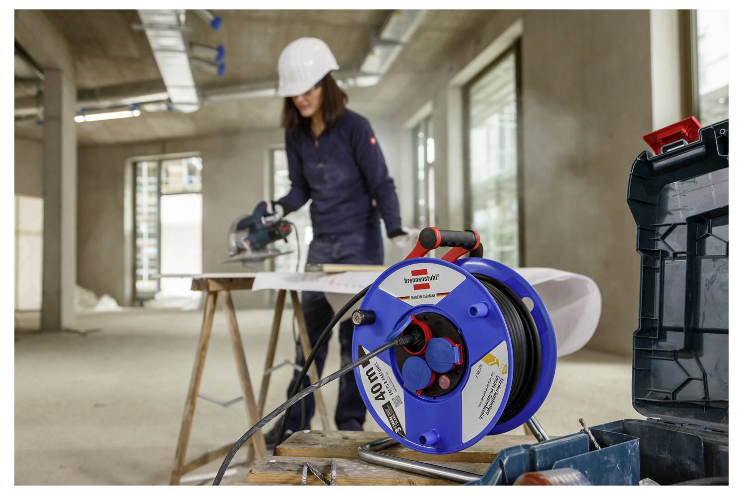 Construction worker in a hard hat uses a circular saw on a wooden plank, with a blue cable reel and toolbox in the foreground at an unfinished building site.