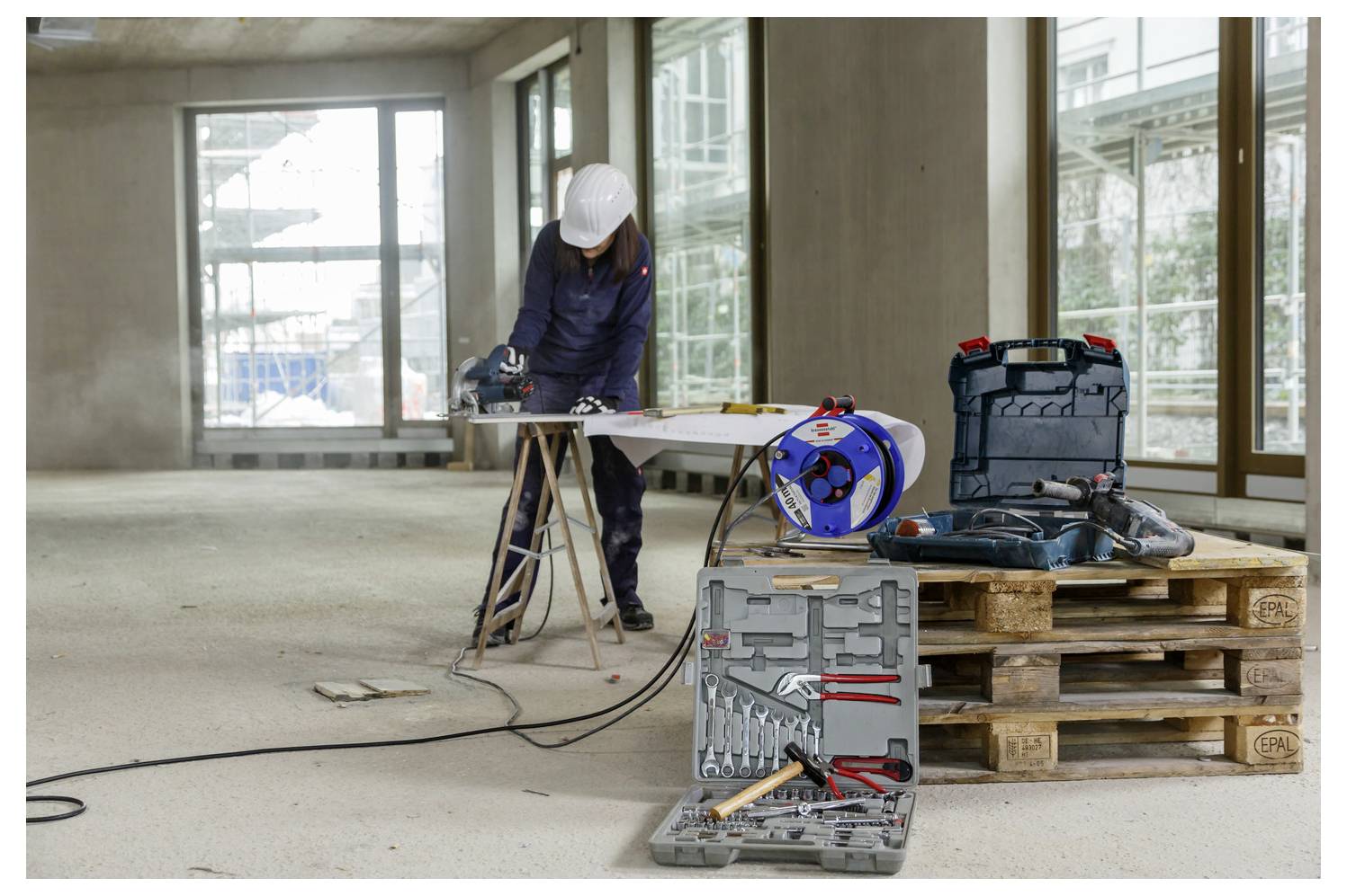 Construction worker in a hard hat using a power saw inside an unfinished room, with tools and materials on pallets in the foreground.