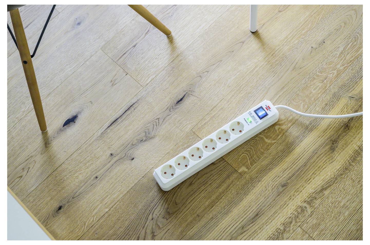 Power strip with six outlets on a wooden floor, plugged in and switched on under a table.