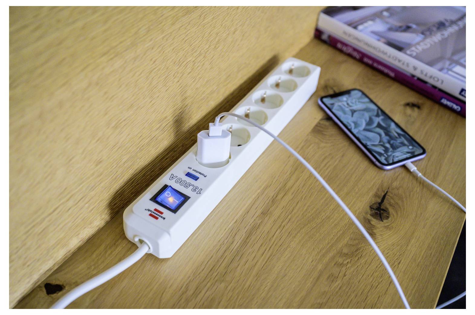 Power strip with a phone charger plugged in on a wooden desk, next to a smartphone and books, showing device charging setup.
