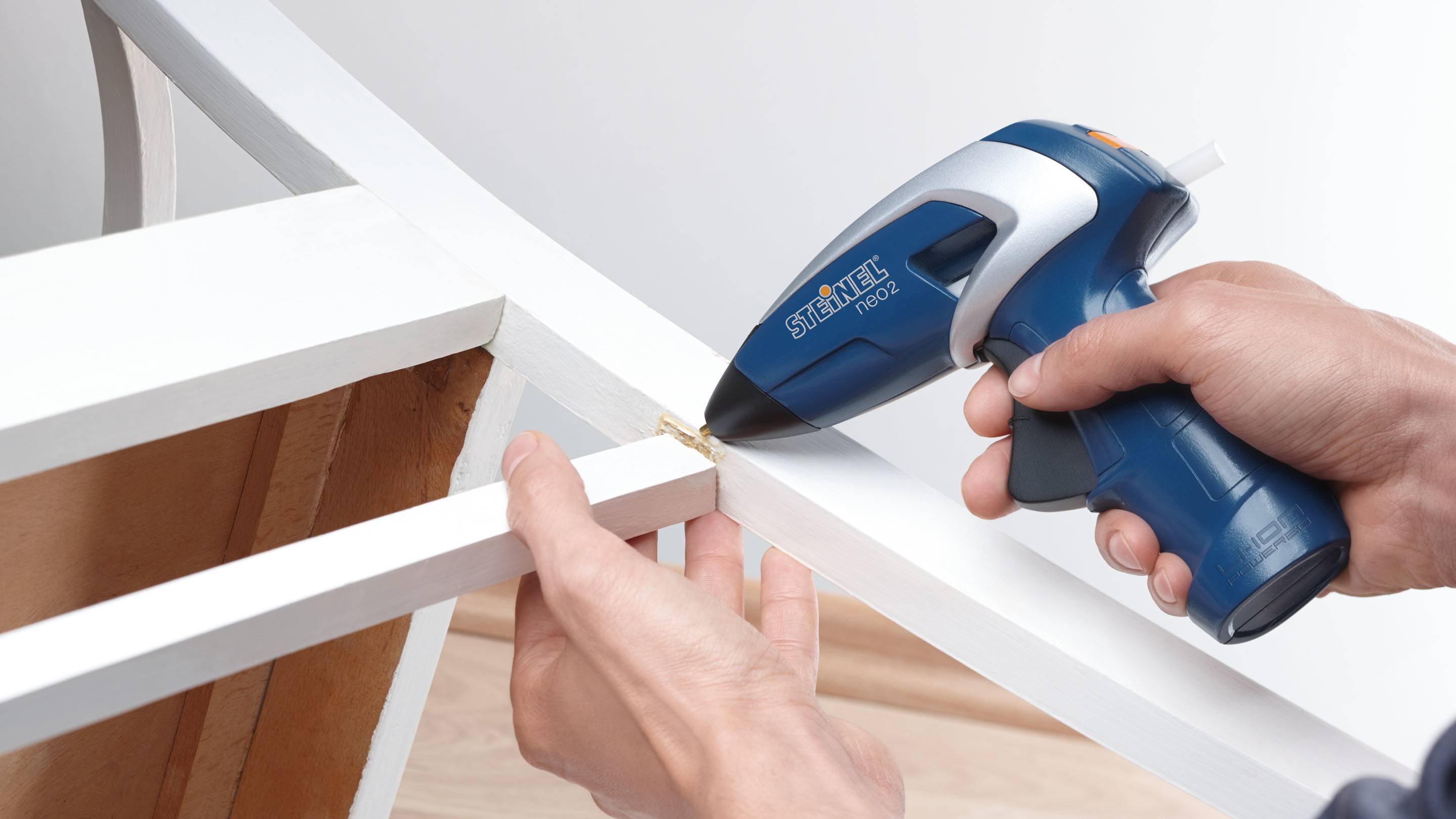 A person is holding a blue hot glue gun and sticking two white wooden pieces together. Craftwork, woodworking.