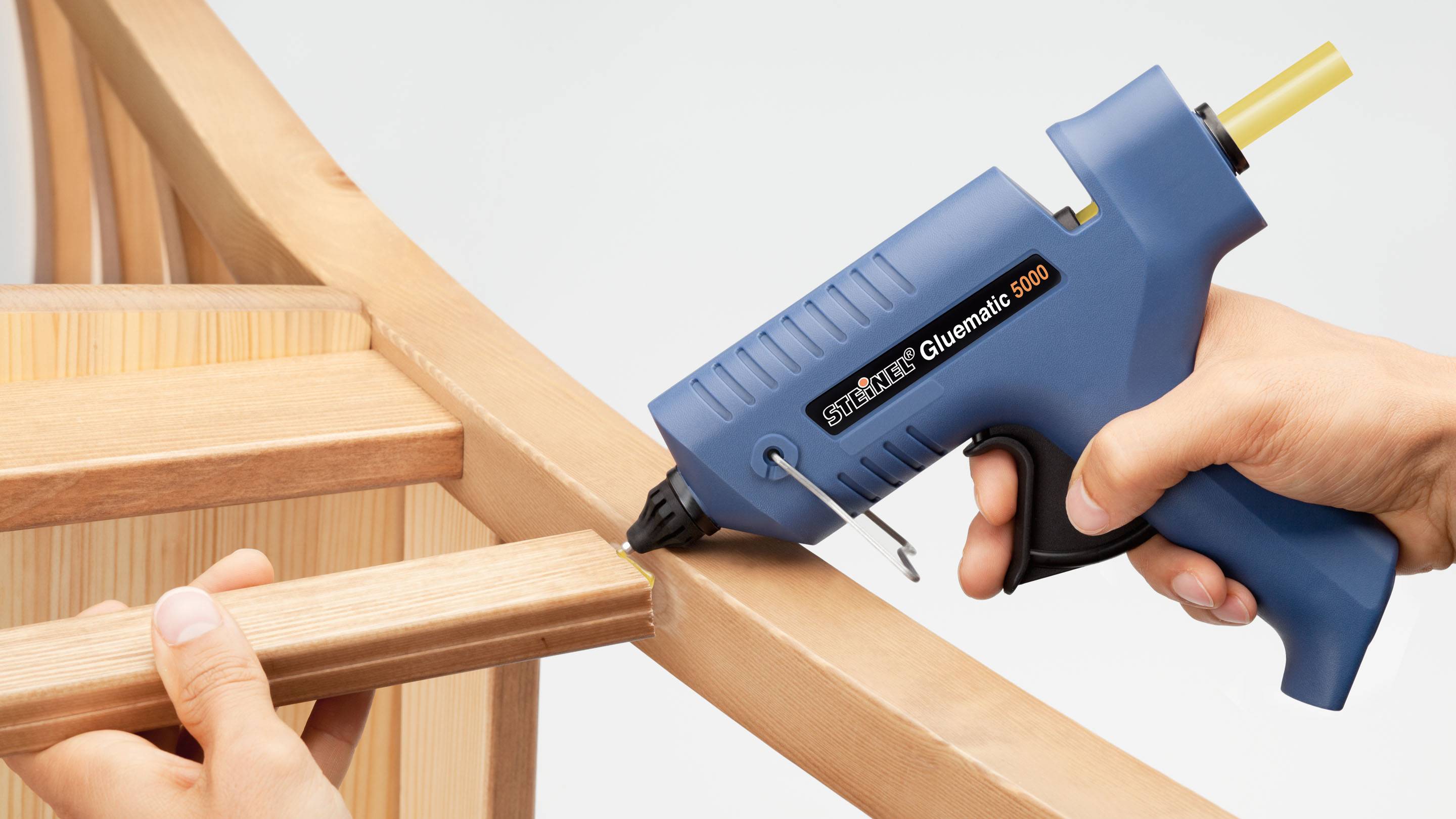 A person is using a hot glue gun to join wooden parts of a piece of furniture. A hand is holding and guiding the device close to the joint.