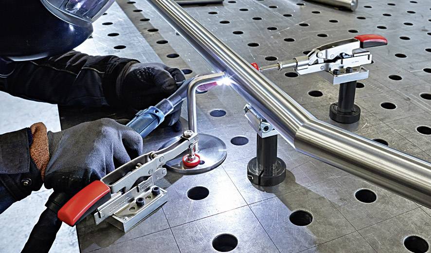 'Person welding metal pipe on perforated table wearing safety glasses and gloves. Clamps are holding the pipe stable.'