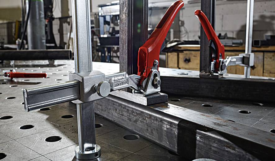 Two red quick-release clamps and an adjustable metal stop on a workbench prepared for welding work.