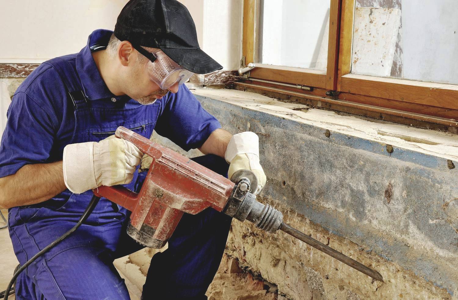 A tradesman in blue workwear is using a large drill hammer to renovate an old wall next to a window.