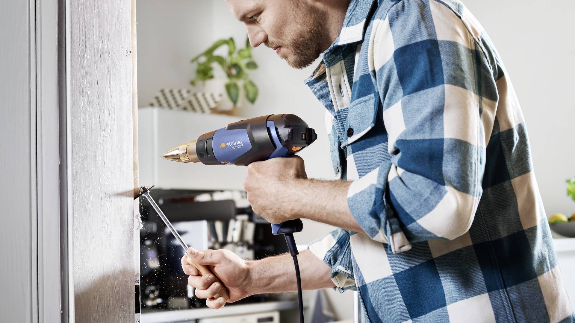 A man in a checked shirt is using a drill to bore a hole into a piece of wood. Plants can be seen in the background.