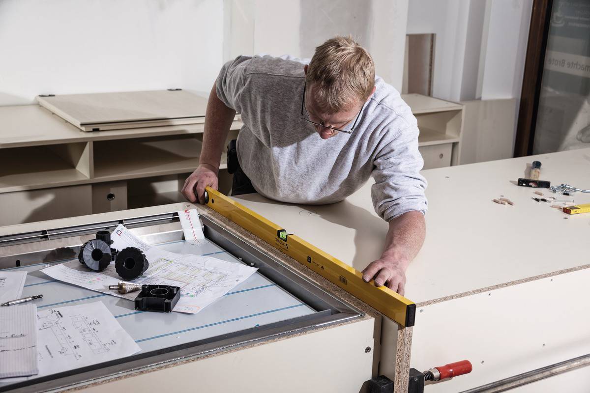 A man is measuring a wooden board with a spirit level. Building plans and tools are lying on the table, indicating a construction project.