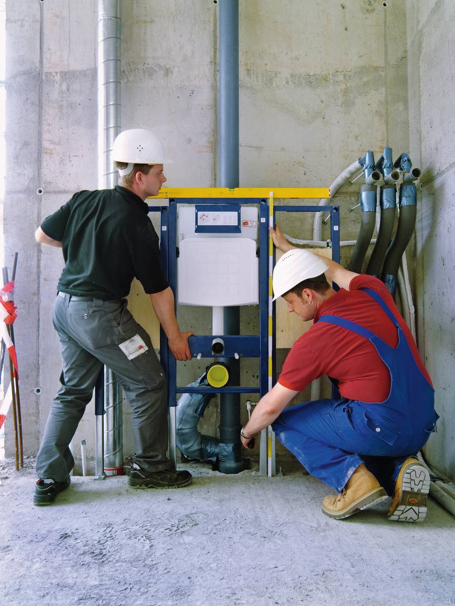 Two construction workers wearing white hard hats are installing a pre-wall element for a toilet in an unfinished building. One is checking the frame, while the other is crouching on the floor.