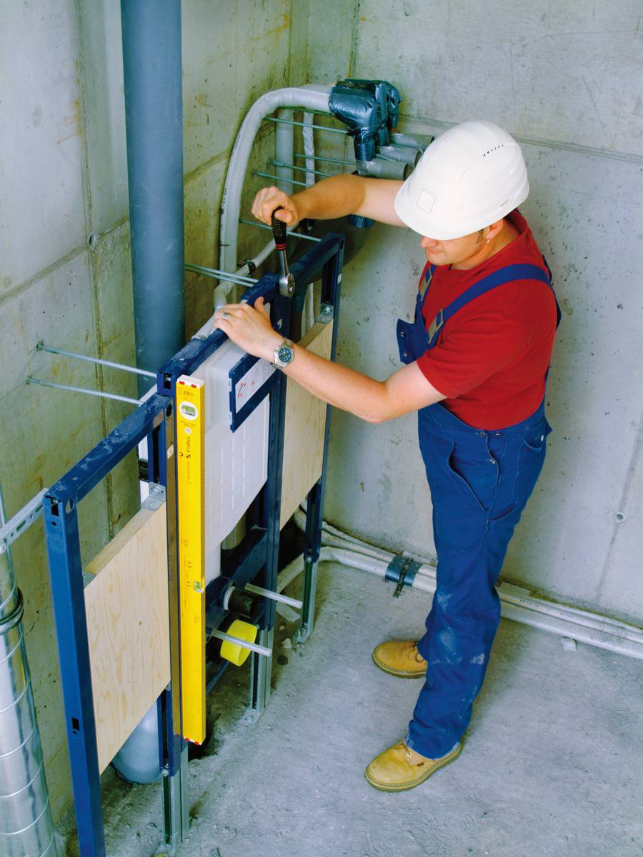 A construction worker wearing a hard hat and red shirt is adjusting a metal structure in an unfinished building, using a spirit level and a hammer.