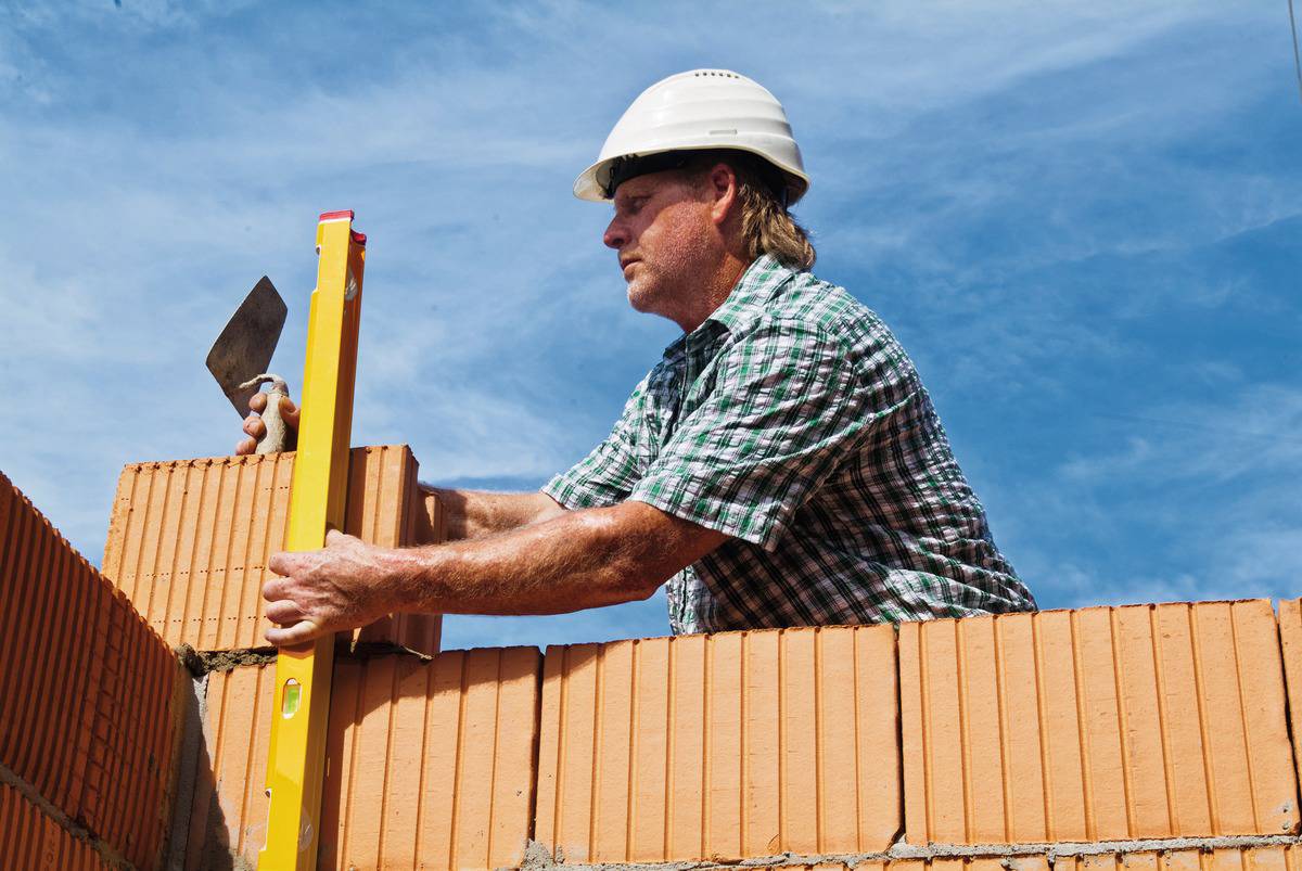 A construction worker wearing a hard hat is aligning brickwork using a spirit level. Clear sky in the background.