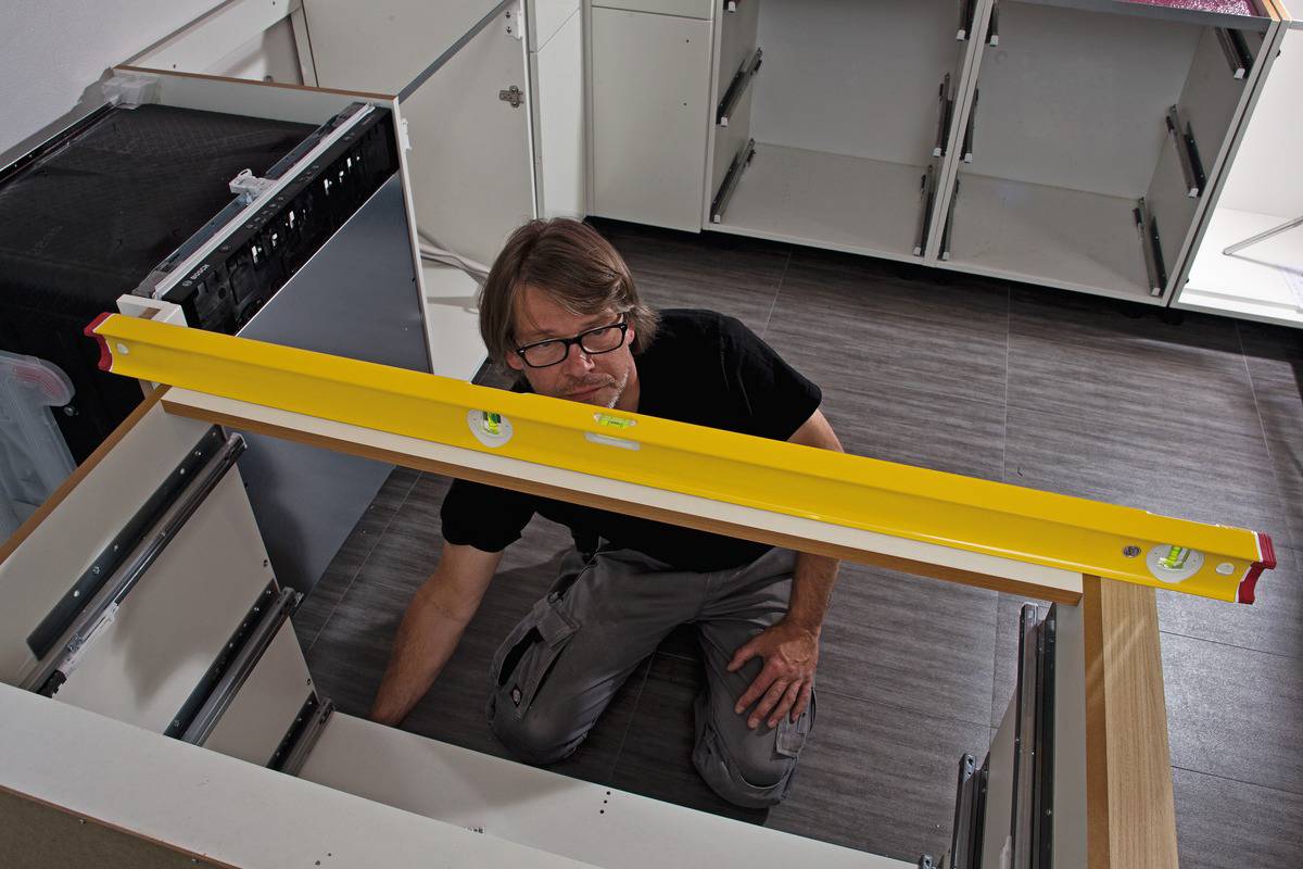 A person is aligning a kitchen cupboard using a spirit level. The room has a dark grey tiled floor and partially installed kitchen units.