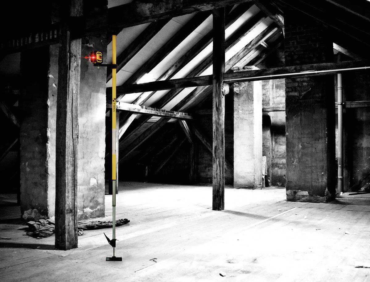 An empty, old attic with exposed wooden beams and pillars. A measuring tape and spirit level are in the foreground.