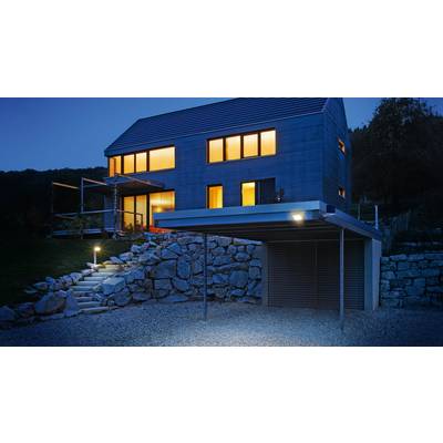 Modern house with large windows and blue facade lighting at night, with stone steps and a carport in the foreground.