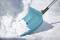 A blue snow shovel clears fresh snow from a frozen pavement. It depicts a wintry scene with a clear sky.