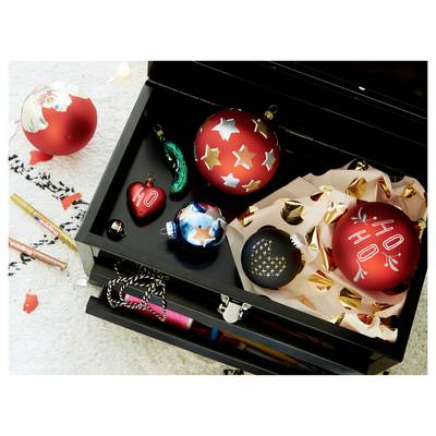 Open black gift box filled with decorated Christmas ornaments, including red, blue, and gold baubles, with wrapping paper and pens nearby.
