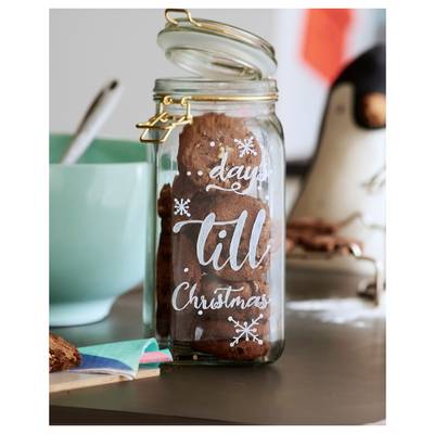 Glass jar of cookies labeled 'days till Christmas' on a kitchen counter, with a teal bowl and blurred holiday decorations in the background.