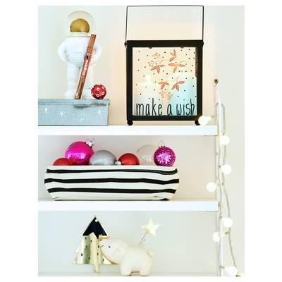 A decorated white shelf with a 'make a wish' sign, toy astronaut, striped basket of ornaments, and small holiday figurines with string lights.