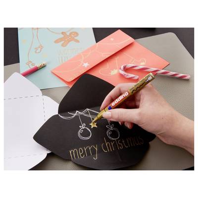 Hand decorating a black Christmas card with a gold pen, writing 'merry christmas' and drawing ornaments on a table with other cards and a candy cane.