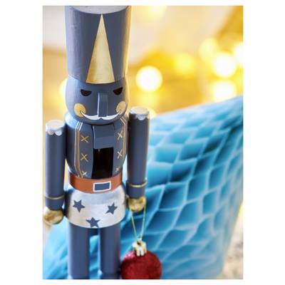 Toy nutcracker figurine standing on a blue quilted surface, holding a small red ornament, with warm blurred lights in the background.