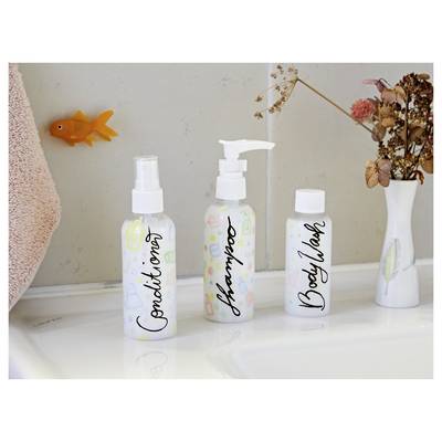 Three labeled white toiletry bottles on a bathroom counter, including 'Conditioner', 'Shampoo', and 'Body Wash', beside a vase of dried flowers and an orange fish decoration.