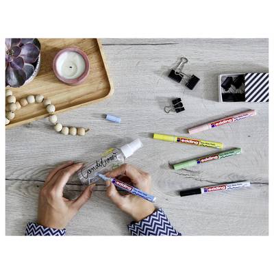 Hands decorating a glass bottle with the word 'traditional' using a blue paint marker on a gray tabletop with craft supplies nearby<br>