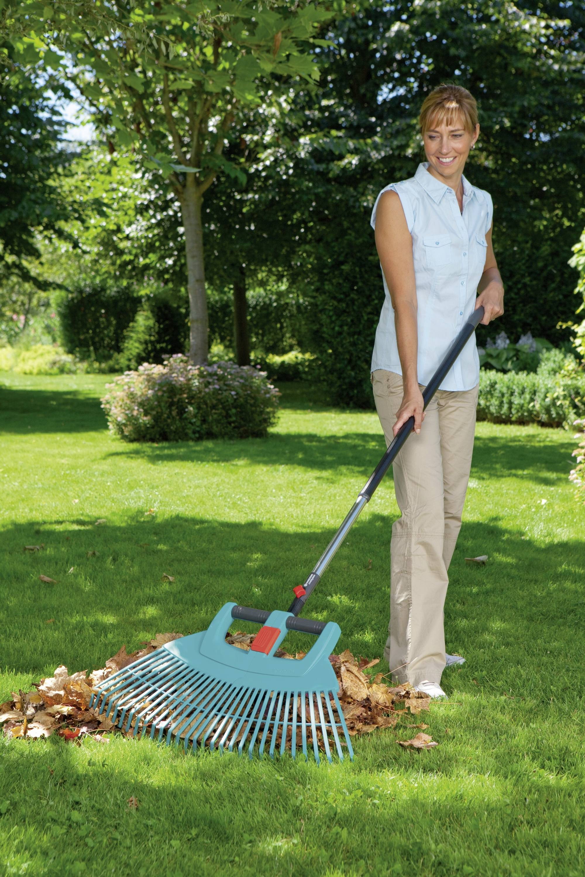 A woman is raking leaves on a green lawn in a garden with trees in the background. She is smiling and wearing a sleeveless blouse.