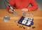 A person in a red shirt is engraving a small metal box with a hand tool. Small parts and a tool set are lying on the table.
