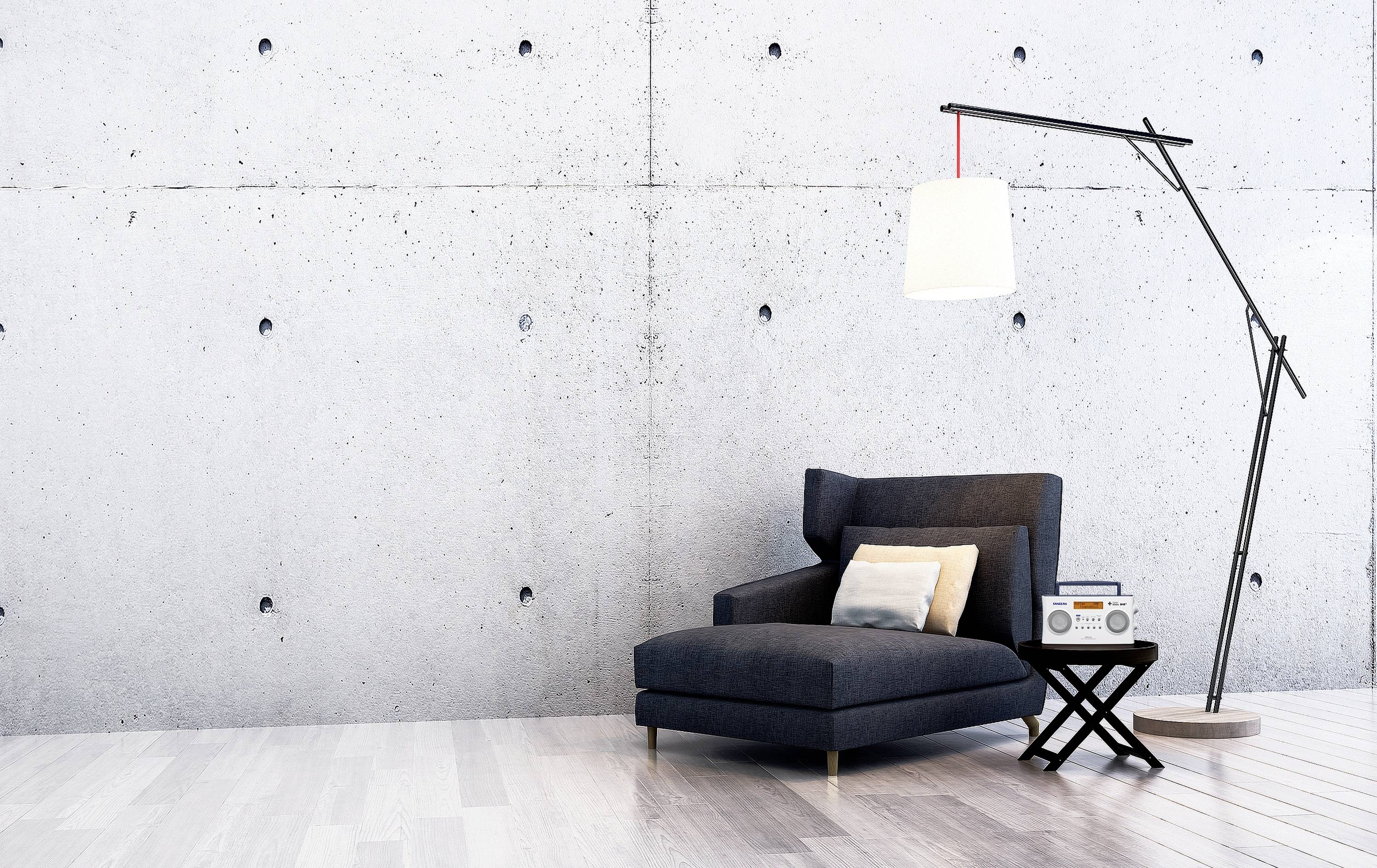 A modern, minimalist space with a grey armchair, a standard lamp, and a small table with a radio in front of a concrete wall.
