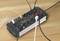 A black surge-protected power strip with six sockets is lying on a wooden table. Several cables are plugged in.