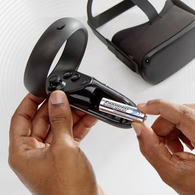 A person is holding a VR controller remote and inserting an AA battery. A VR headset is visible in the background.