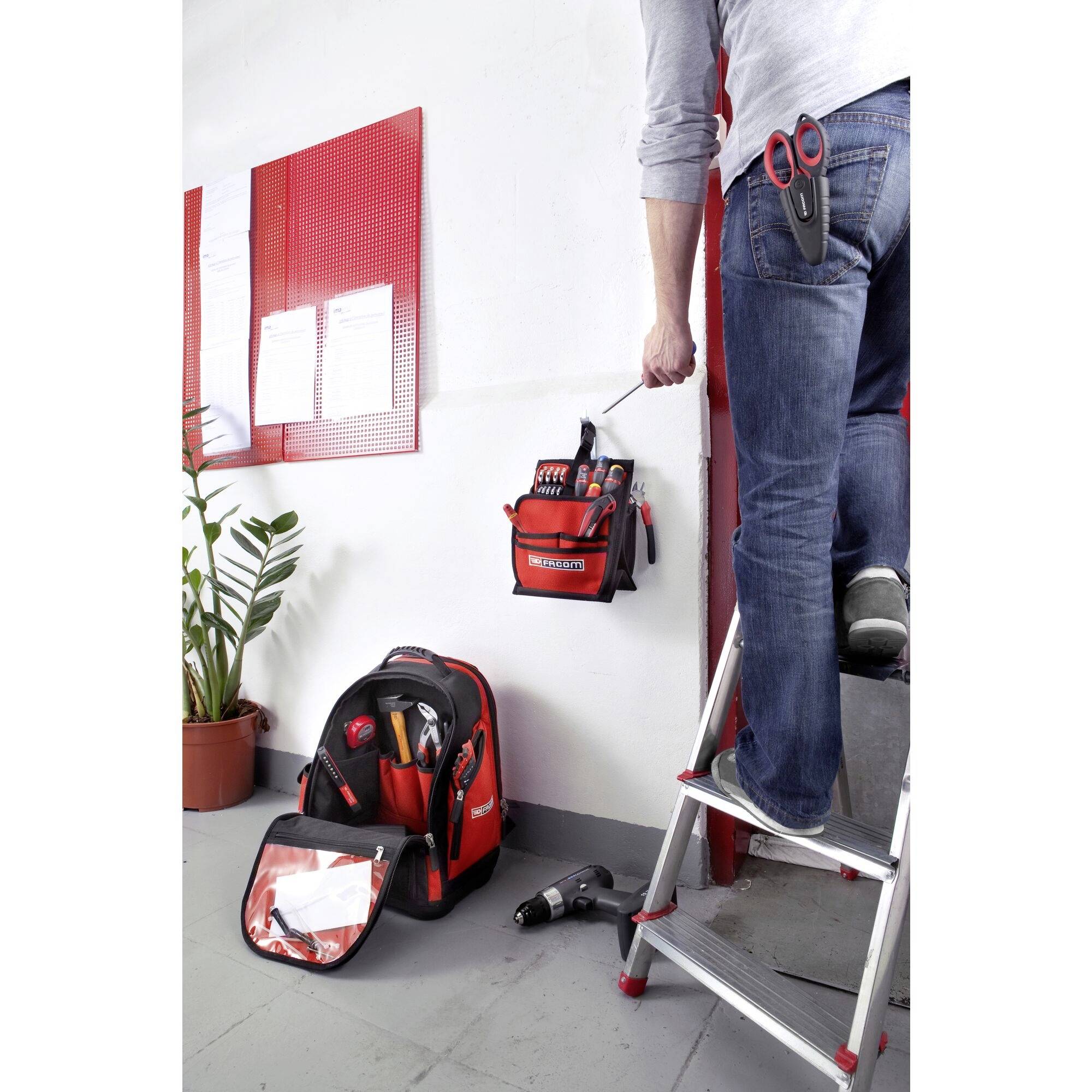A person is standing on a ladder and reaching into a tool bag hanging on the wall. A ring binder and a plant are standing on the floor.