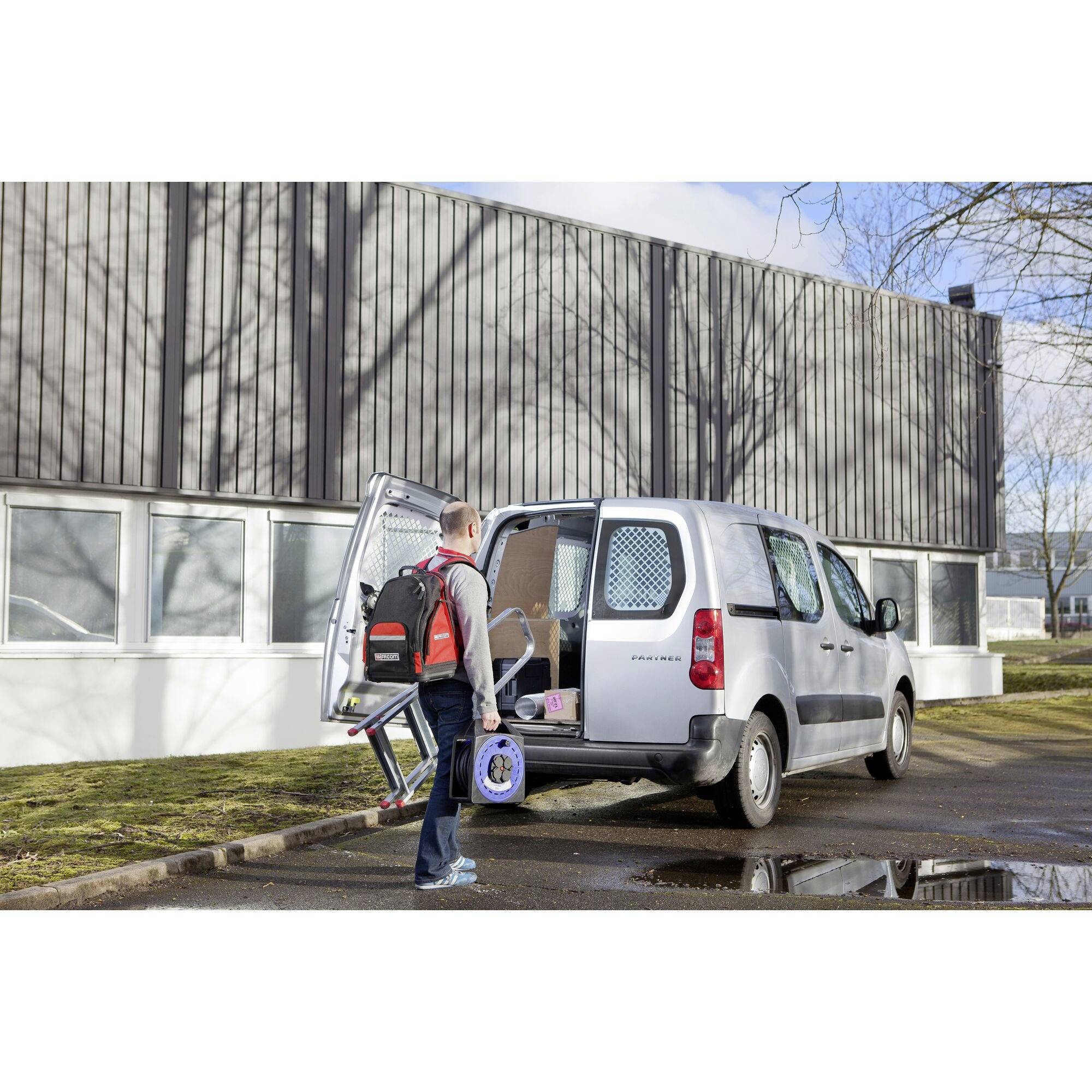 A person is lifting a wheelchair to the rear of a silver delivery van in front of a grey building.