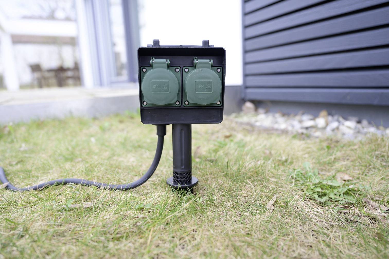 'Black outdoor socket with two green covers, mounted on lawn. A modern house can be seen in the background.'