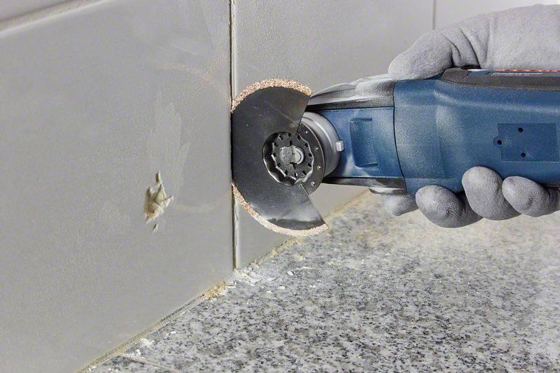 'Someone is using an oscillating tool with a circular blade to cut tiles on the wall while wearing gloves.'