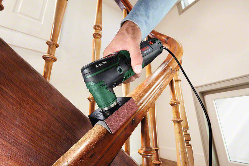 A person is sanding a wooden handrail with an electric sanding tool. The tool has a rectangular sanding head.