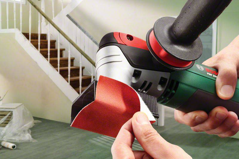 A person is attaching sandpaper to a sanding machine in a renovated room with a staircase in the background.