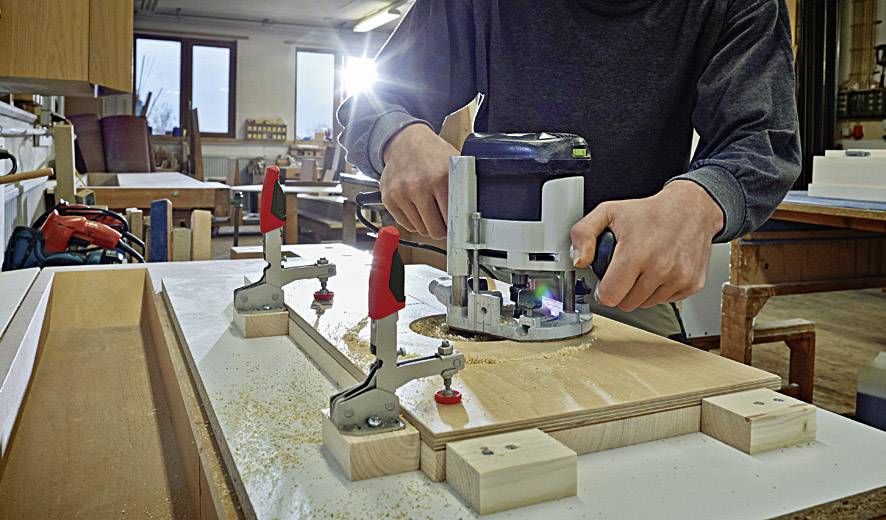 A person is using a router to work on wood on a workbench in a workshop. Wooden pieces are secured with clamps.