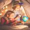 Two children are playing under a blanket. One is holding a book and a torch, the other is wearing a hat and examining a globe.