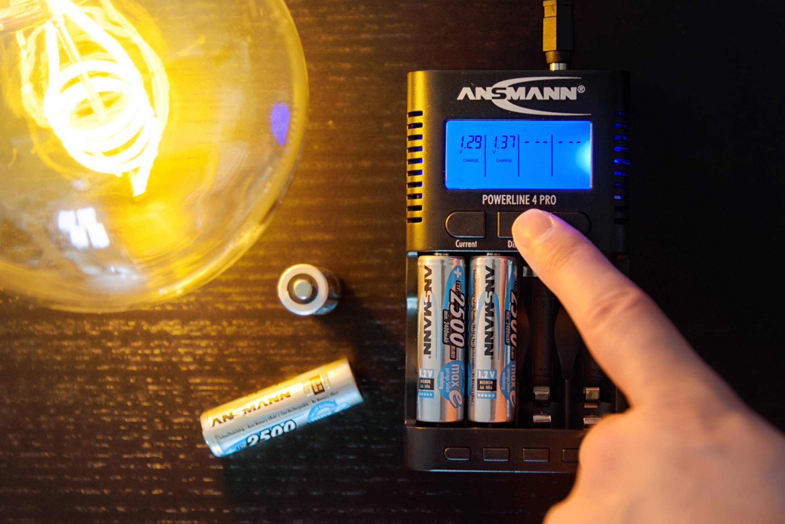 A person touches a button on a battery charger next to charging AA batteries. A light bulb glows in the background.
