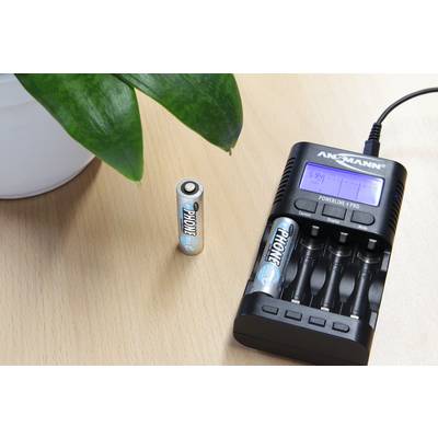 A charger is charging four AA batteries on a wooden table. One battery is standing beside them. A part of a green plant is visible in the top left corner.