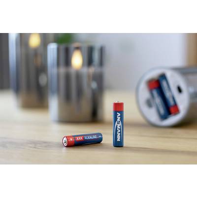 Two red and blue AAA batteries are sitting on a table; a shiny candle and an open tin of batteries are blurred in the background.