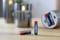 Two red and blue AAA batteries are sitting on a table; a shiny candle and an open tin of batteries are blurred in the background.