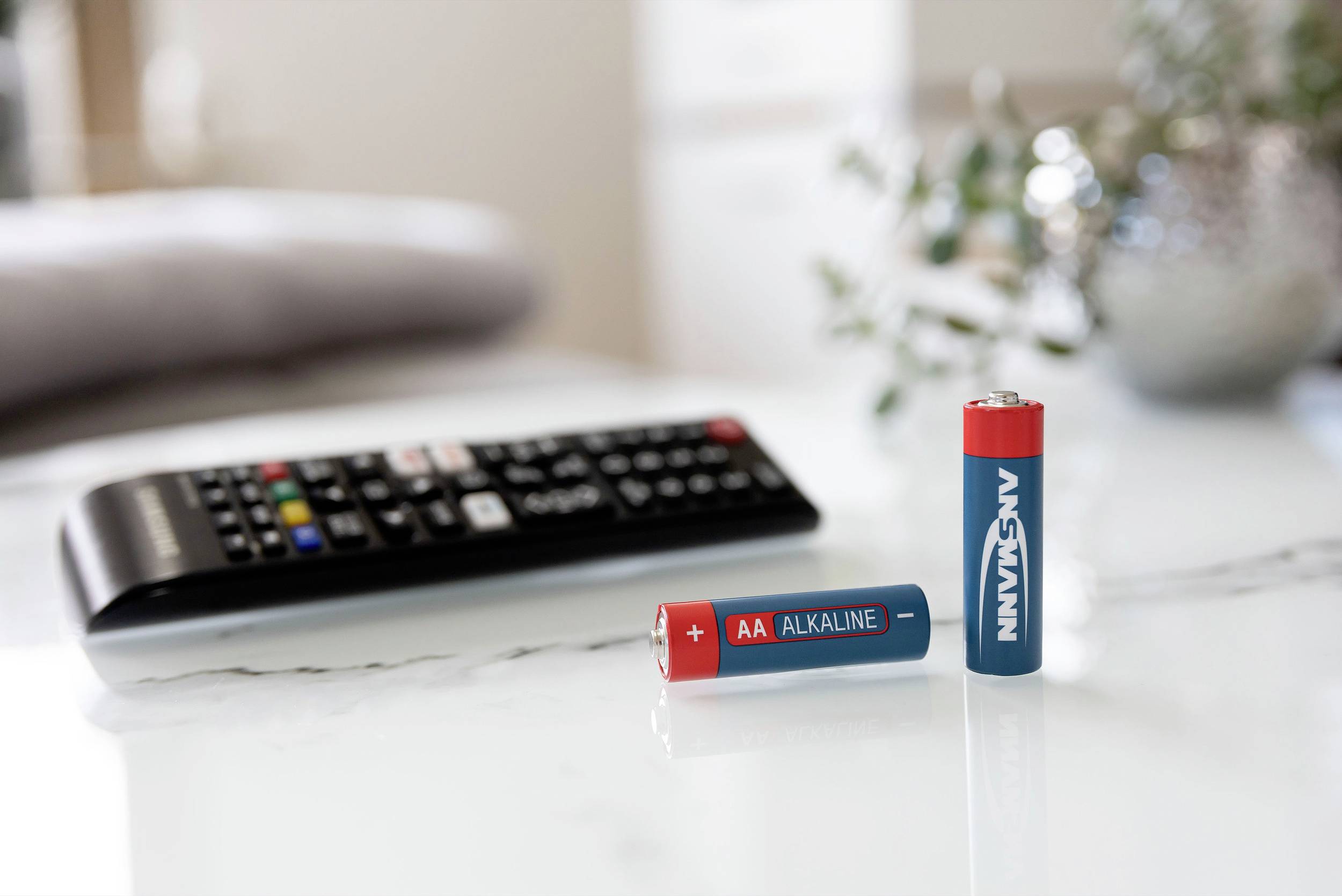 Two AA batteries, one lying down with an 'Alkaline' label, in front of a remote control on a marble table. Background slightly out of focus.