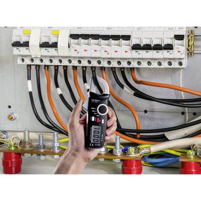 A hand holds a black electrical measuring device in front of an open electrical panel with multiple cables and circuit breakers.